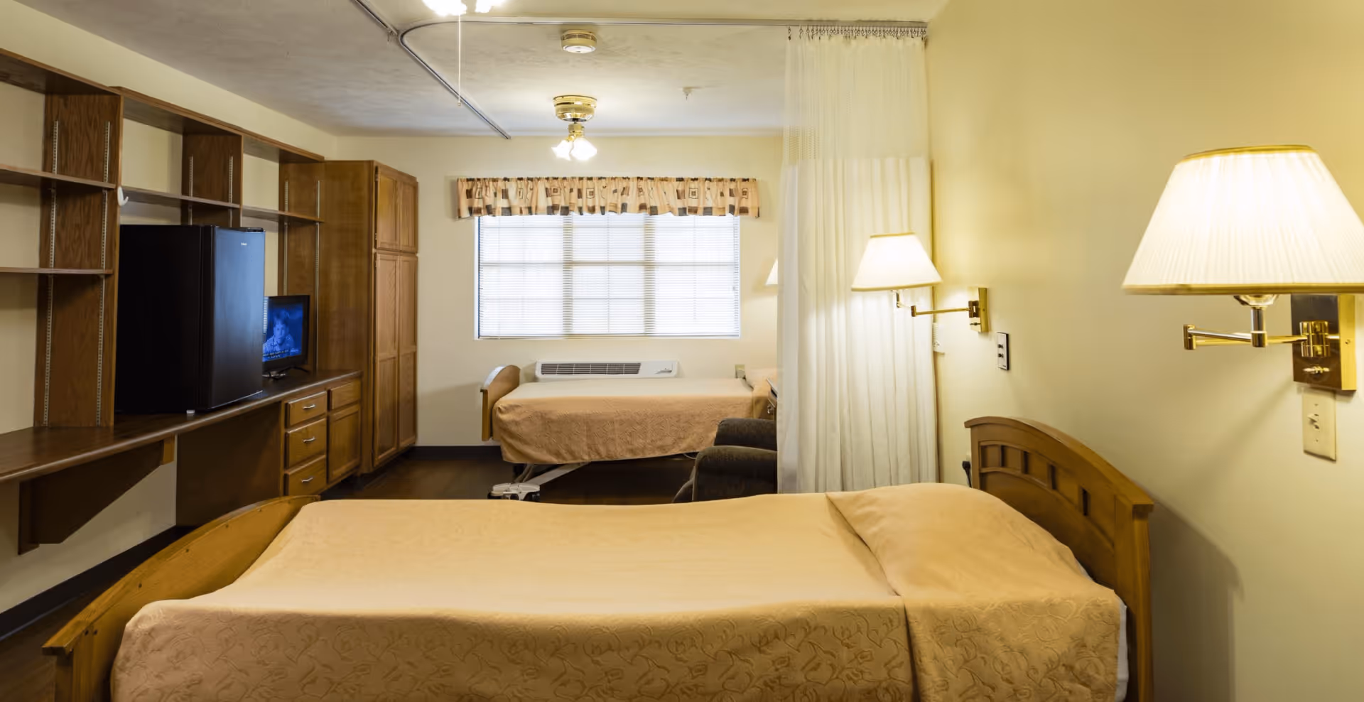 Two-bed senior living resident room with wooden furniture, a TV, bedside lamps, and a window.