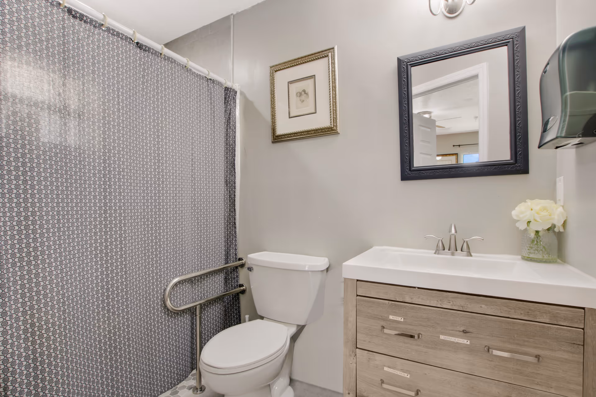 A bathroom with a white toilet, a wooden vanity with a white sink, a black-framed mirror above the sink, a gray patterned shower curtain, a metal grab bar next to the toilet, a small framed picture on the wall, and a vase with white flowers on the vanity.