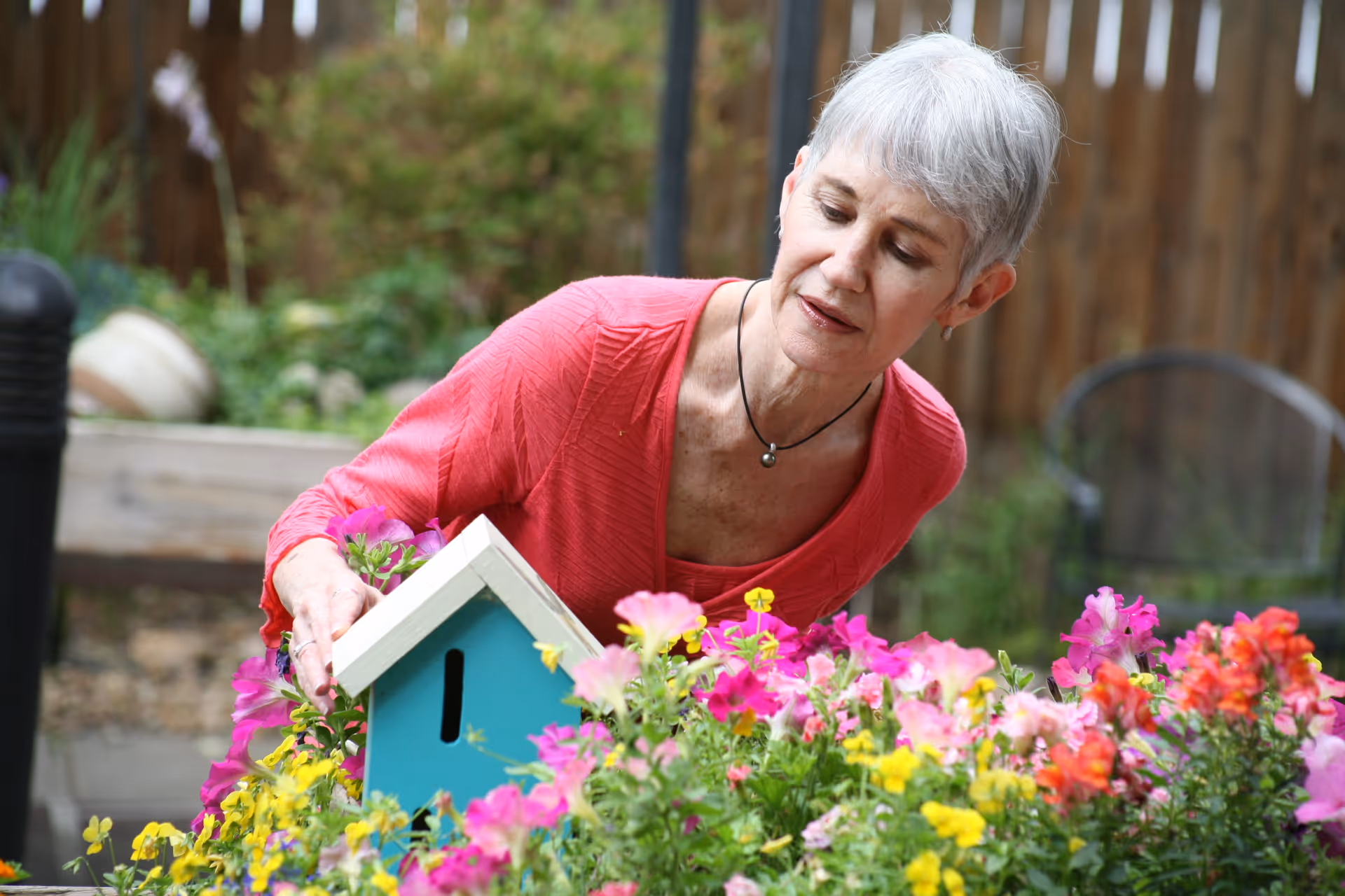An elderly woman with short gray hair wearing a red top is tending to a garden filled with colorful flowers. She is placing or adjusting a small blue birdhouse among the flowers. The background shows a wooden fence and some garden furniture.