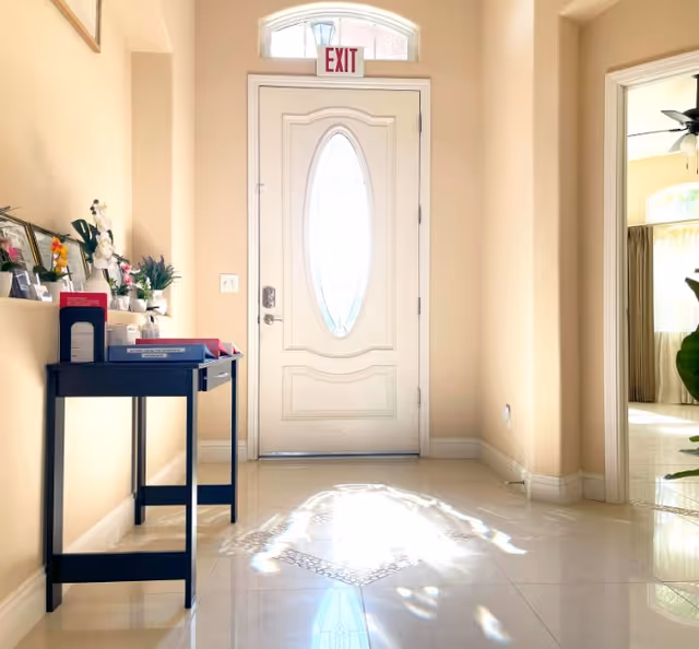 Bright entryway with a white front door featuring an oval glass panel and an exit sign above it. A black console table on the left holds various decorative items including framed photos, flowers, and books. The floor is shiny and reflects light coming through the door. To the right, an open doorway leads to a room with a ceiling fan and large windows with curtains.