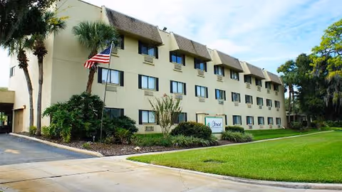 Three-story beige senior living building with windows, manicured lawn, palms, and an American flag.