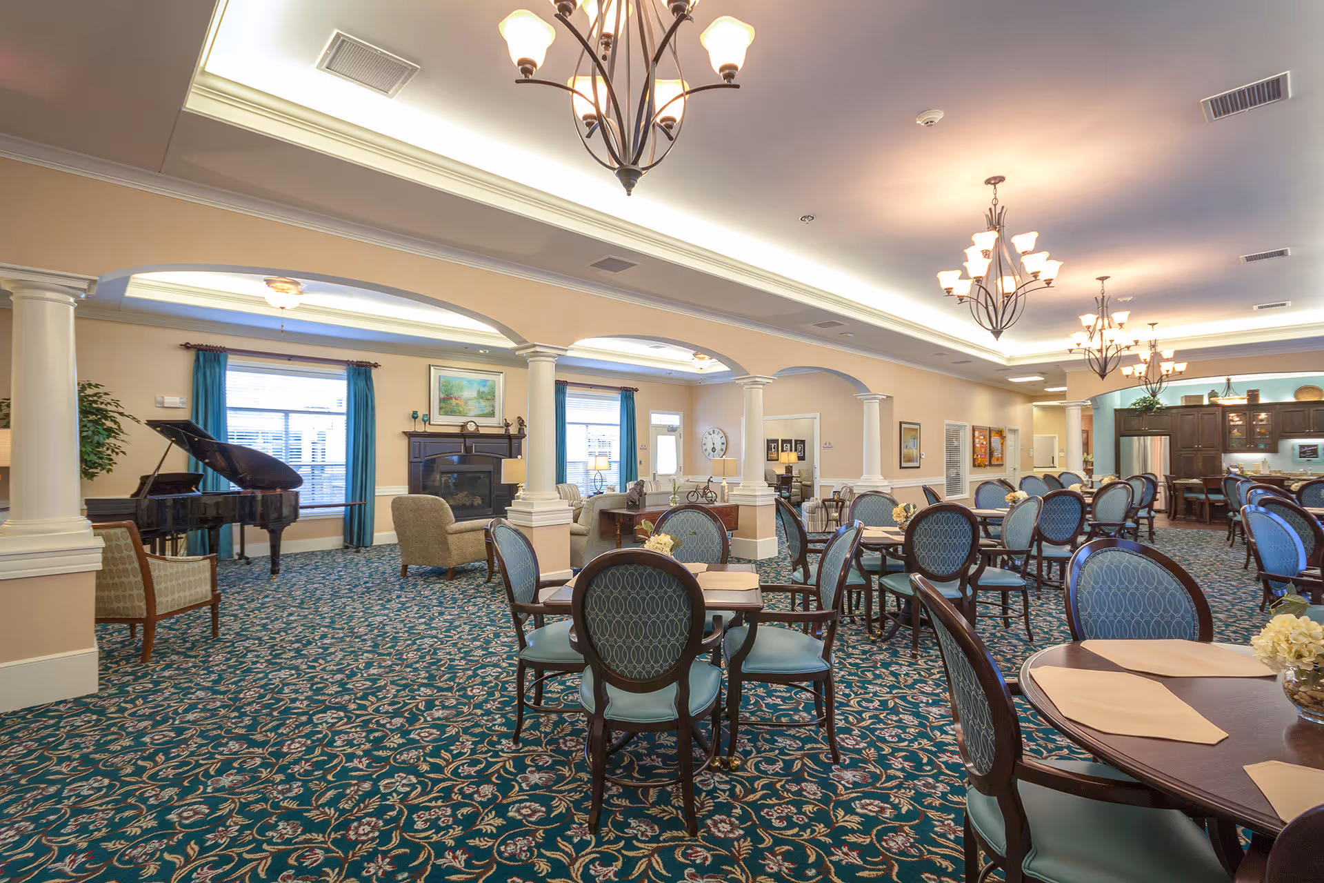 A spacious and elegant common area in a senior living facility featuring multiple round tables with blue upholstered chairs, a patterned carpet, and a seating area with armchairs near a fireplace and a grand piano. The room is well-lit with chandeliers and has beige walls with white columns and blue curtains on the windows.