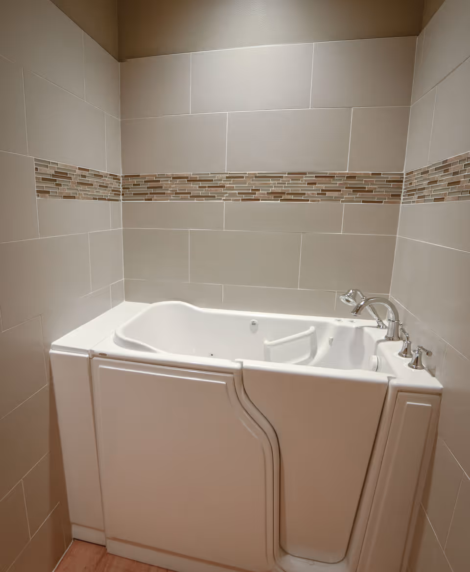 A walk-in bathtub with a door for easy access, surrounded by beige tiled walls with a decorative horizontal strip of small rectangular tiles in various shades of brown and beige.