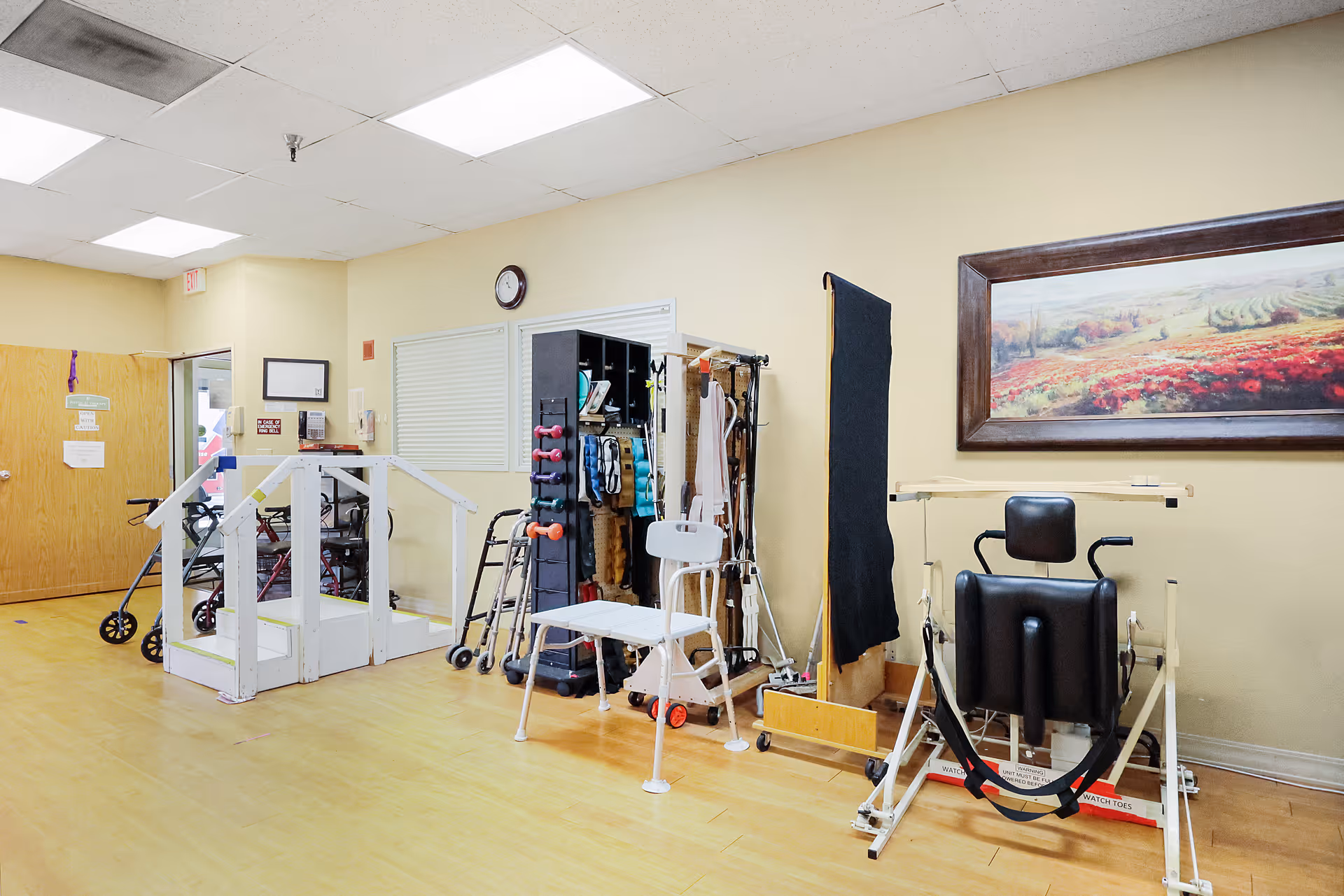 A therapy/rehab room with walkers, exercise equipment, parallel bars, and a framed landscape painting on the wall.