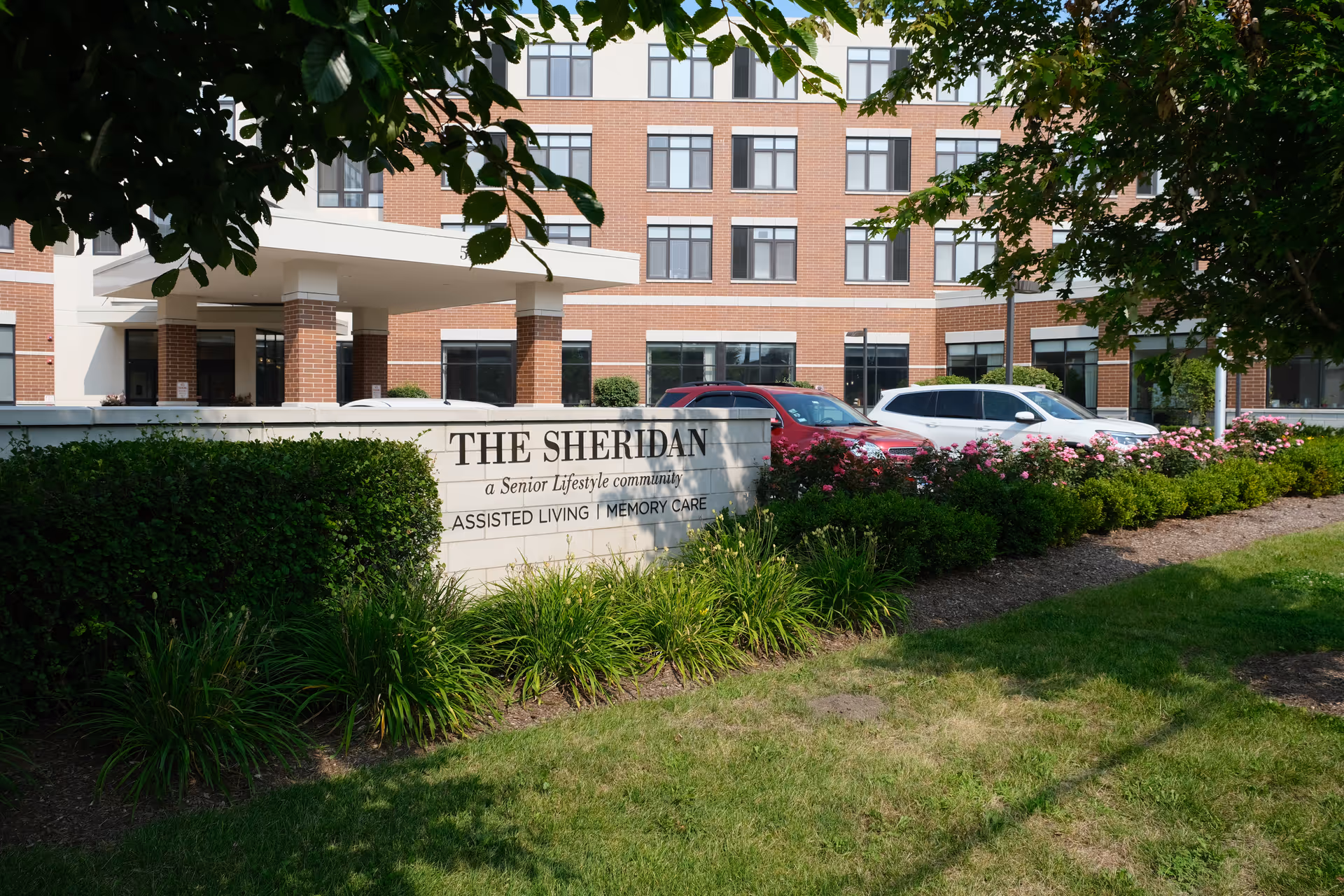 Exterior view of The Sheridan at Park Ridge senior living facility showing the building's brick facade, entrance canopy, parked cars, and landscaped garden with green bushes and pink flowers in front of a sign that reads 'THE SHERIDAN a Senior Lifestyle community ASSISTED LIVING | MEMORY CARE'.