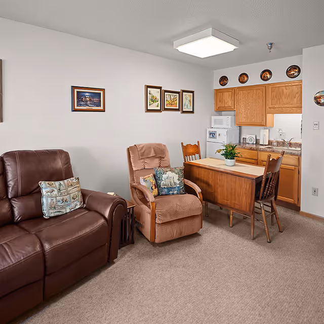 A cozy living area with a brown leather recliner and a beige fabric recliner, each with decorative pillows. A small wooden dining table with two chairs is positioned near a kitchenette with wooden cabinets, a microwave, and a refrigerator. The walls are decorated with framed pictures and decorative plates above the cabinets.