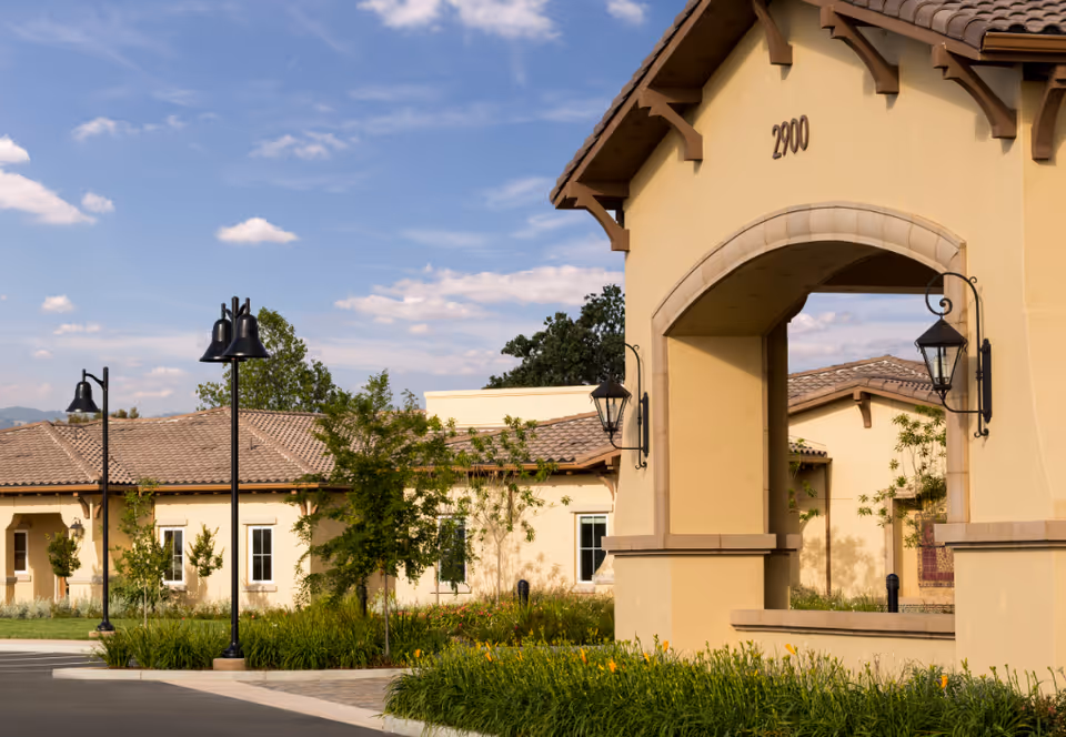 Exterior view of a beige stucco building with a tiled roof and arched entryway numbered 2000, surrounded by landscaped greenery and street lamps under a partly cloudy blue sky.