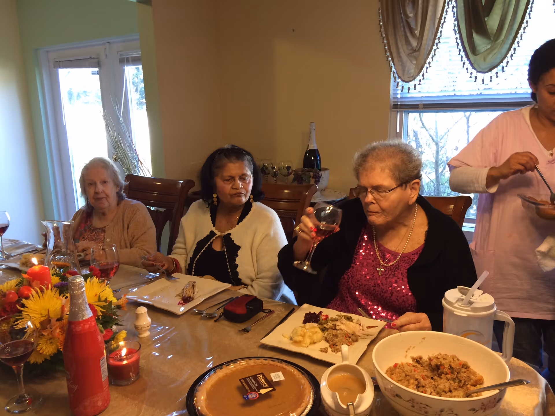Three elderly women sitting at a dining table with plates of food and drinks, one woman holding a glass of red beverage. A caregiver in a pink uniform is serving food on the right side. The table is decorated with flowers, candles, and a pumpkin pie.