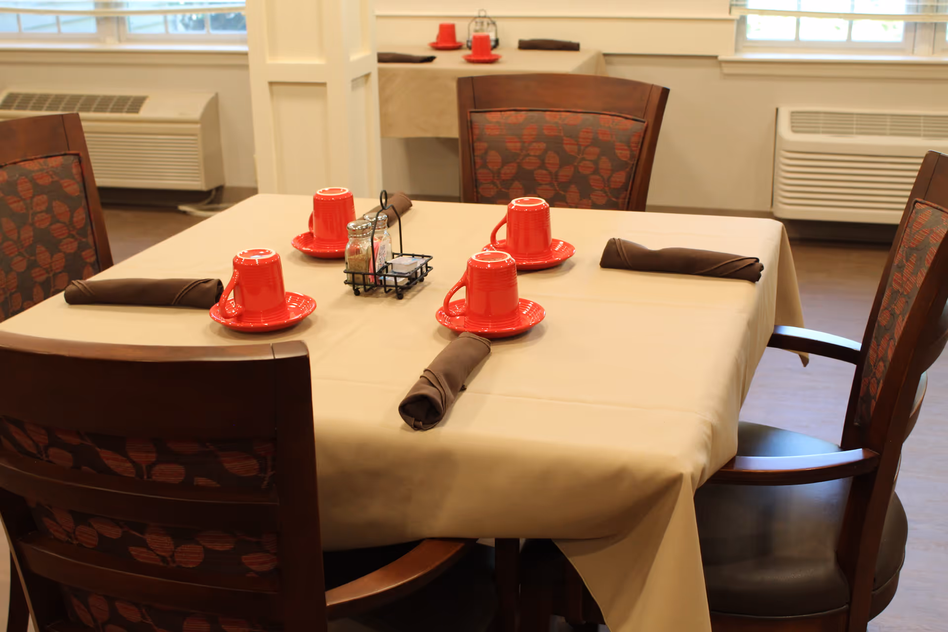 A dining table set for four with a beige tablecloth, four red cups placed upside down on matching red saucers, and four brown rolled napkins. The table is surrounded by wooden chairs with patterned upholstery. In the background, there is another table similarly set and windows letting in natural light.