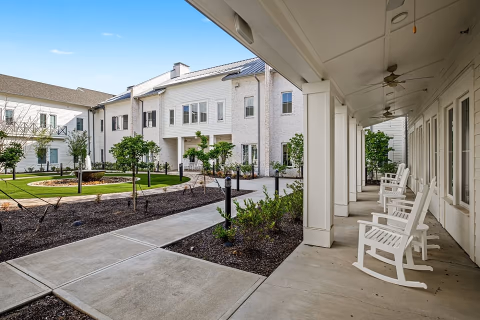 Outdoor courtyard area at The Claiborne at Shoe Creek featuring a circular fountain surrounded by grass and small trees, with a covered walkway on the right side lined with white rocking chairs and ceiling fans.