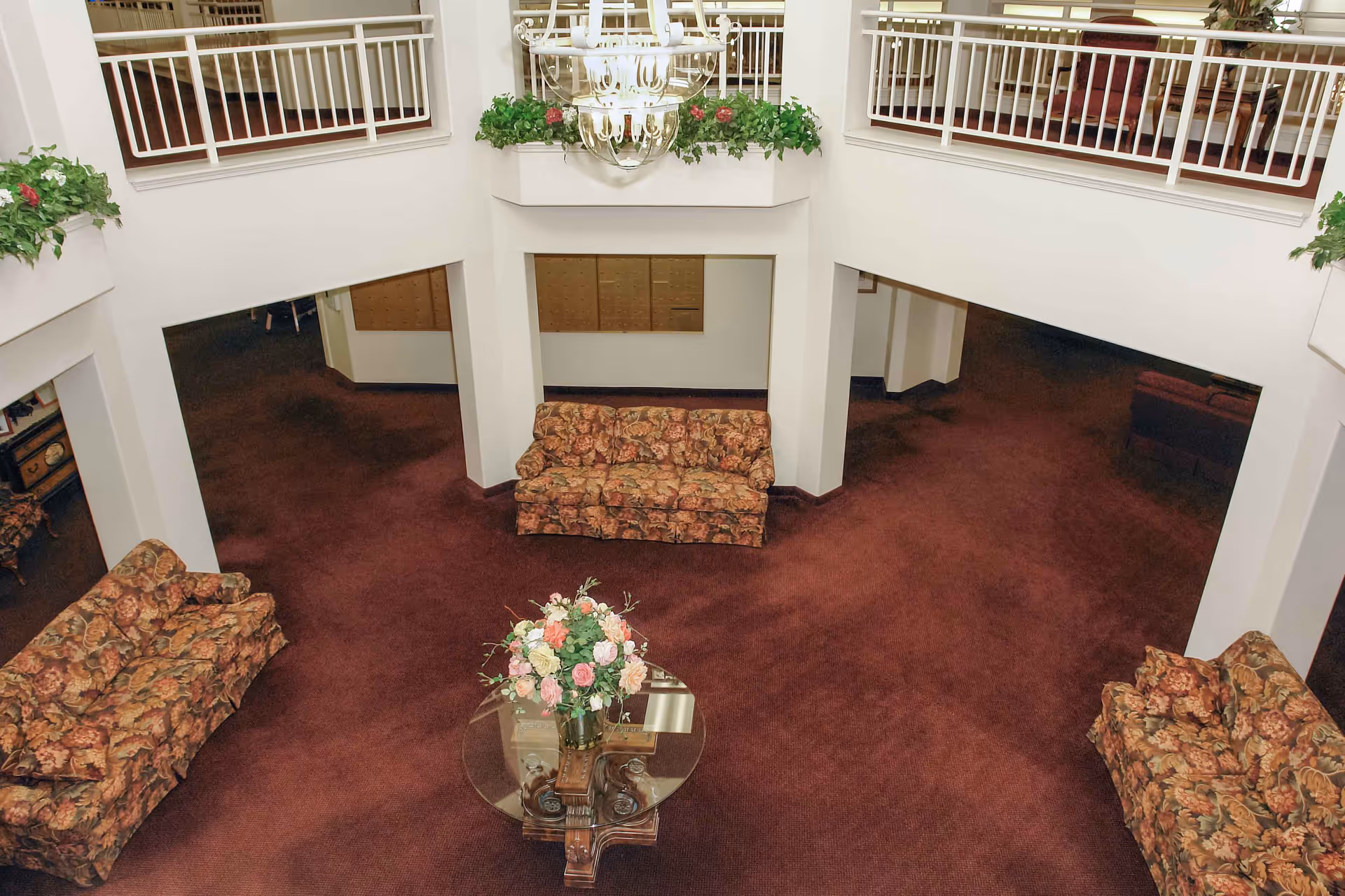 View down into a two-story lobby/atrium with floral upholstered sofas, a glass coffee table topped with flowers, a chandelier, balcony railings, and maroon carpet.