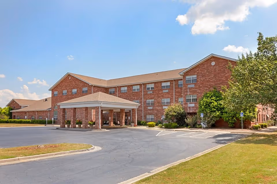 Three-story brick senior living building with a covered entrance and adjacent parking lot under a blue sky.