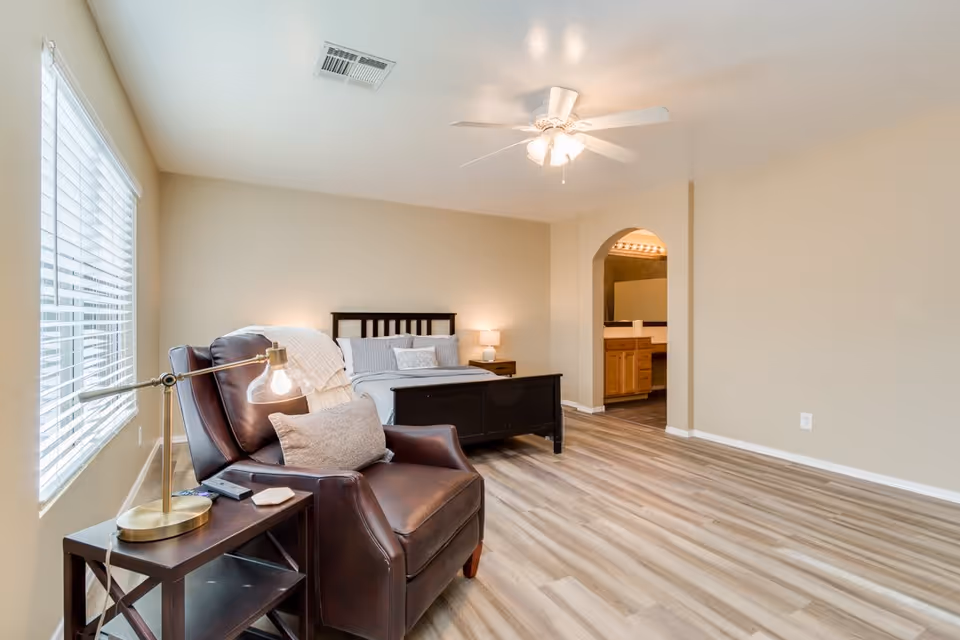 A bright bedroom with a large window on the left, a dark wooden bed with white and gray bedding against the far wall, a brown leather armchair with a side table and lamp in the foreground, and an open doorway leading to a bathroom with a vanity and mirror.