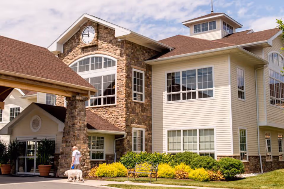 Exterior view of a senior living facility building with stone and beige siding, large windows, a clock above the entrance, and a covered driveway. A person is walking a dog near the entrance, surrounded by green shrubs and yellow flowering plants under a partly cloudy sky.