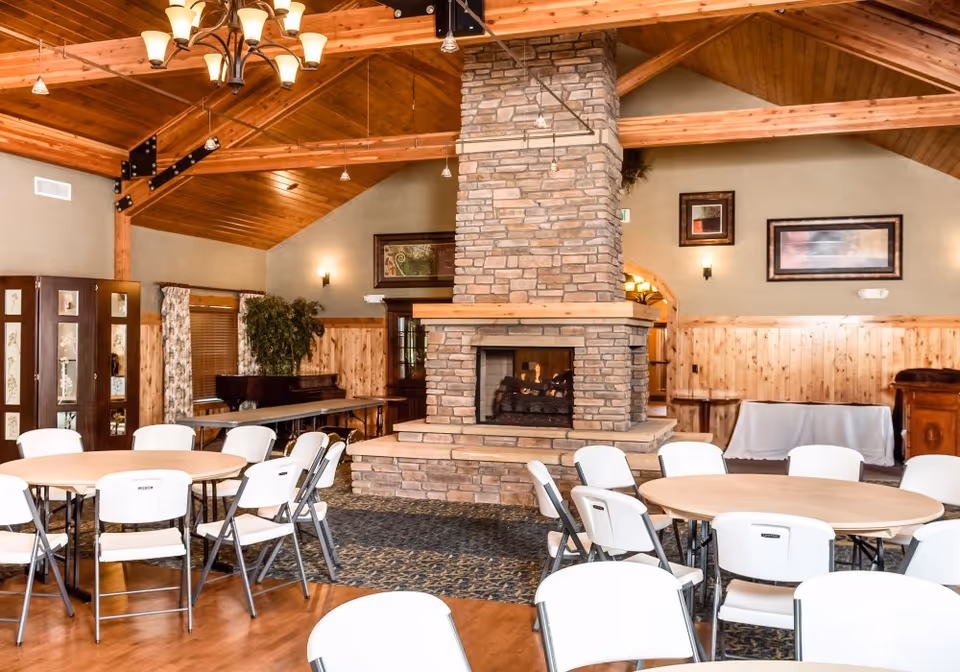 Interior of a senior living facility common area with a large stone fireplace in the center. The room features wooden ceilings with exposed beams, round tables surrounded by white folding chairs, framed artwork on the walls, and a piano in the corner.