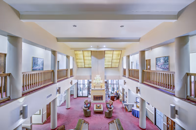 Spacious two-story retirement community lobby with seating around a central fireplace, chandelier, upper balconies, and large skylights.