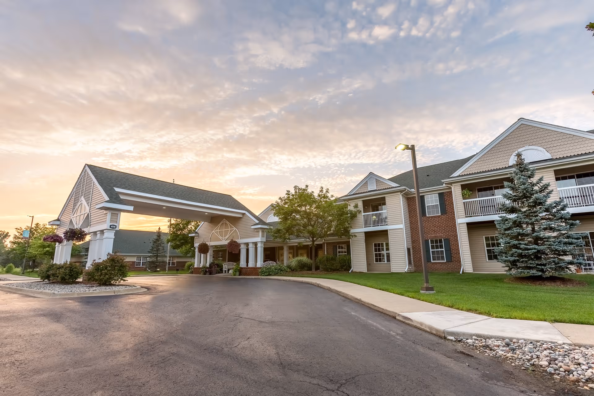 Exterior view of a senior living facility building with a covered entrance, well-maintained lawn, trees, and a paved driveway under a partly cloudy sky during sunset.