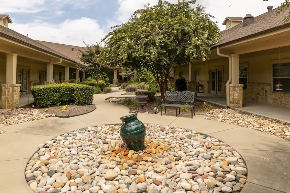 Outdoor courtyard area at Lakeshore Assisted Living and Memory Care featuring a circular rock garden with a green ceramic urn in the center, surrounded by a paved walkway. There are bushes, a tree, and a metal bench along the walkway, with covered walkways and building entrances on both sides.