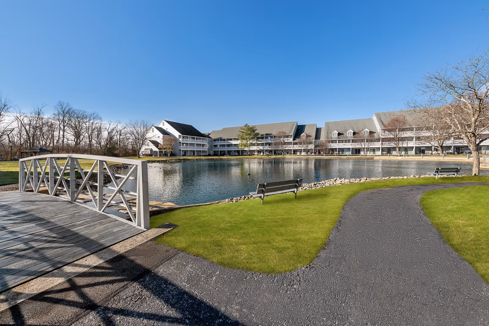 A serene outdoor scene at StoryPoint Cincinnati featuring a small wooden bridge over a pond, a paved walking path, green grass, benches, leafless trees, and a large multi-story building in the background under a clear blue sky.