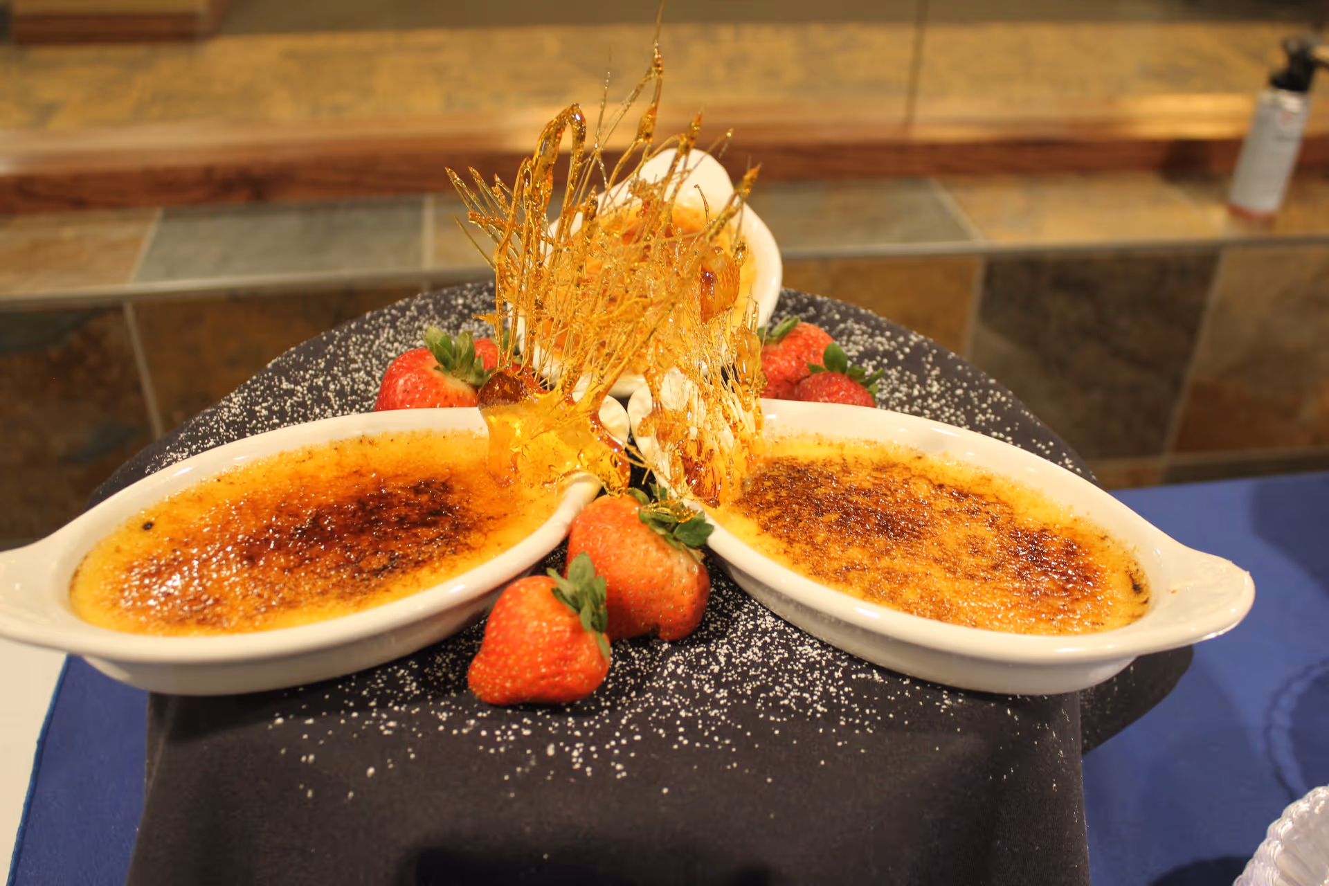 Two dishes of crème brûlée with caramelized sugar tops, garnished with fresh strawberries and an artistic spun sugar decoration, placed on a black cloth with powdered sugar sprinkled around.