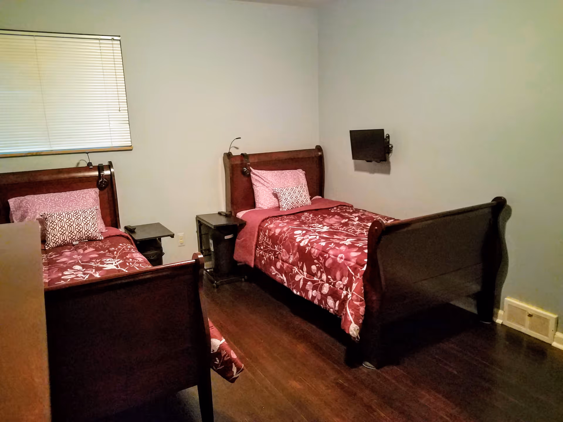 A small bedroom with two single wooden beds placed parallel to each other, each with a pink floral bedspread and pillows. There is a small bedside table between the beds with a trash bin underneath. A small flat-screen TV is mounted on the wall opposite the beds. The room has wooden flooring and a window with closed blinds.