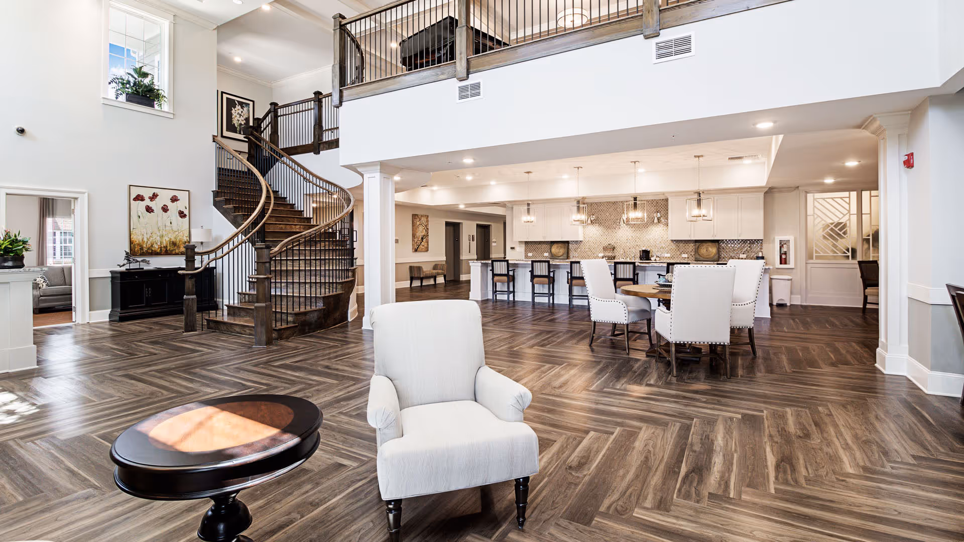 Spacious and bright interior of a senior living facility featuring a modern open floor plan with a curved wooden staircase, a white armchair, a round wooden table with white upholstered chairs, and a kitchen area with bar stools and pendant lighting.