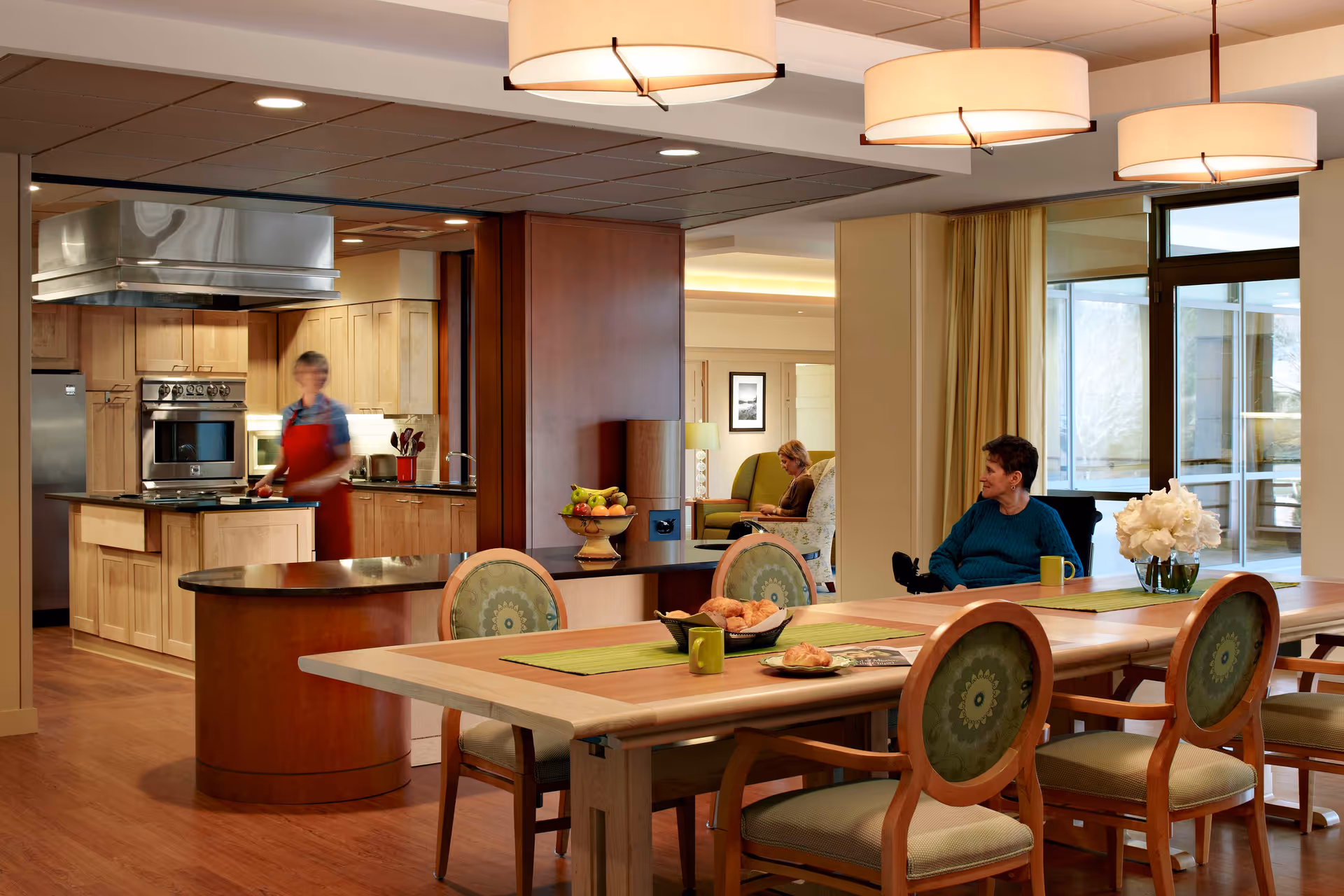 Open communal dining area with a long wooden table and chairs, an adjacent kitchen with a person working, and residents seated nearby.