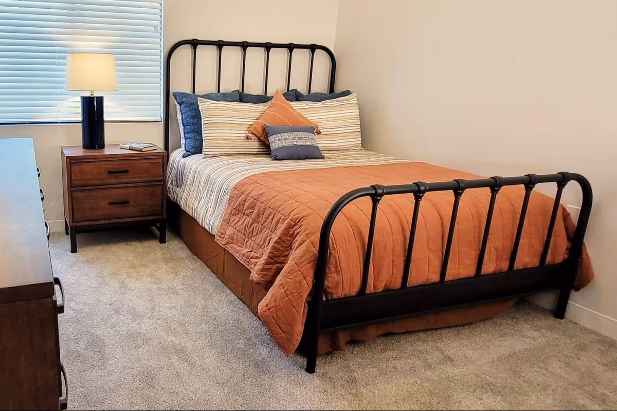 Simple bedroom with a black metal-framed bed dressed in a rust-colored quilt, a wooden nightstand with a lamp, and a window with blinds.