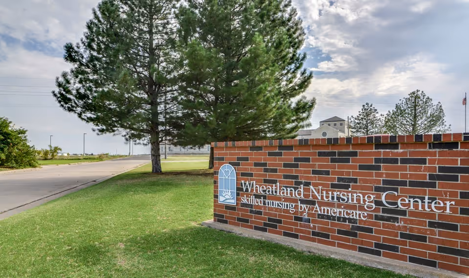 Brick entrance sign reading "Wheatland Nursing Center" on a grassy lawn with large trees and the facility building visible in the background.