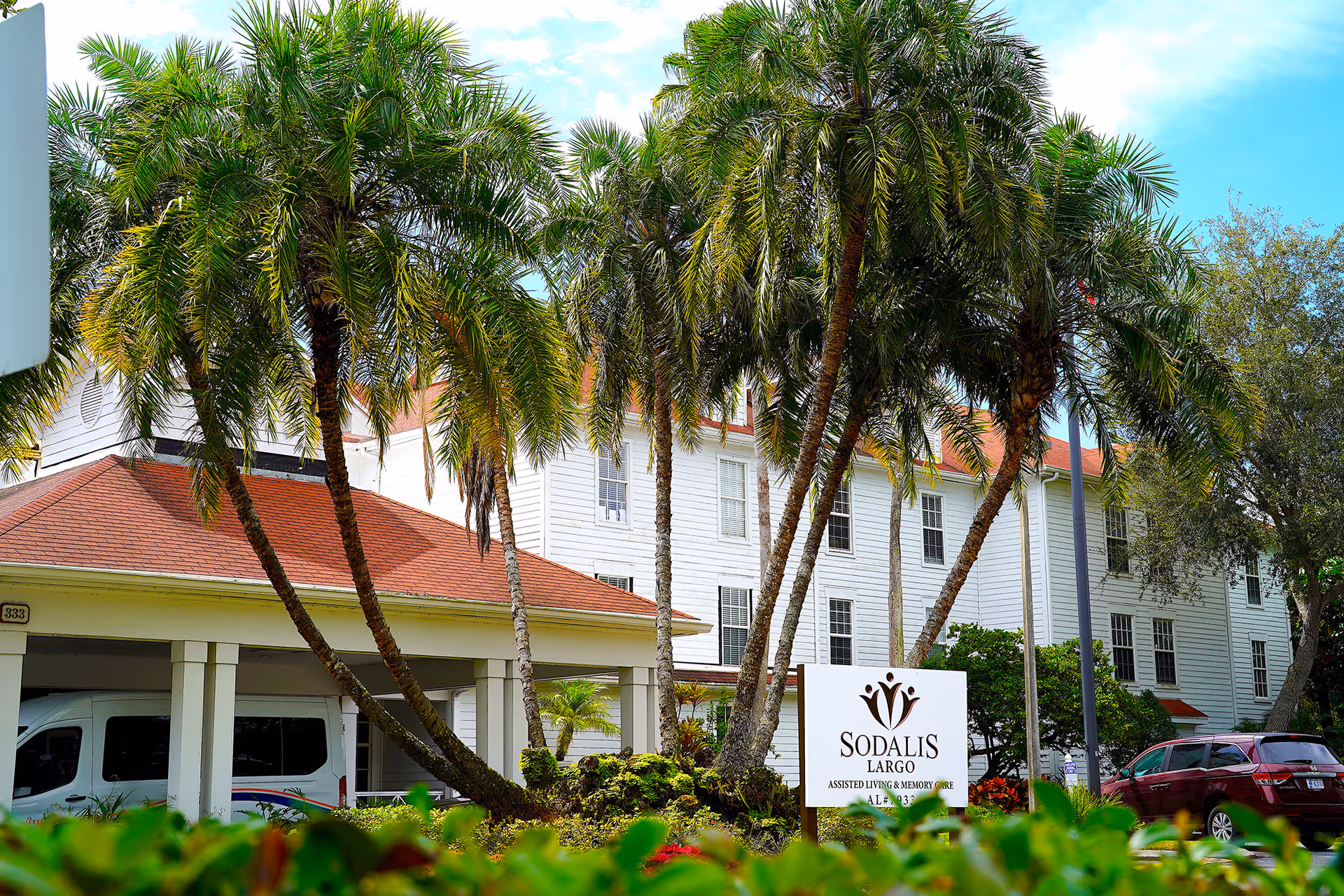 Exterior view of Sodalis of Largo assisted living and memory care facility with palm trees and a white building with red roof in the background. A white sign with the facility's name is visible in the foreground, along with parked vehicles.