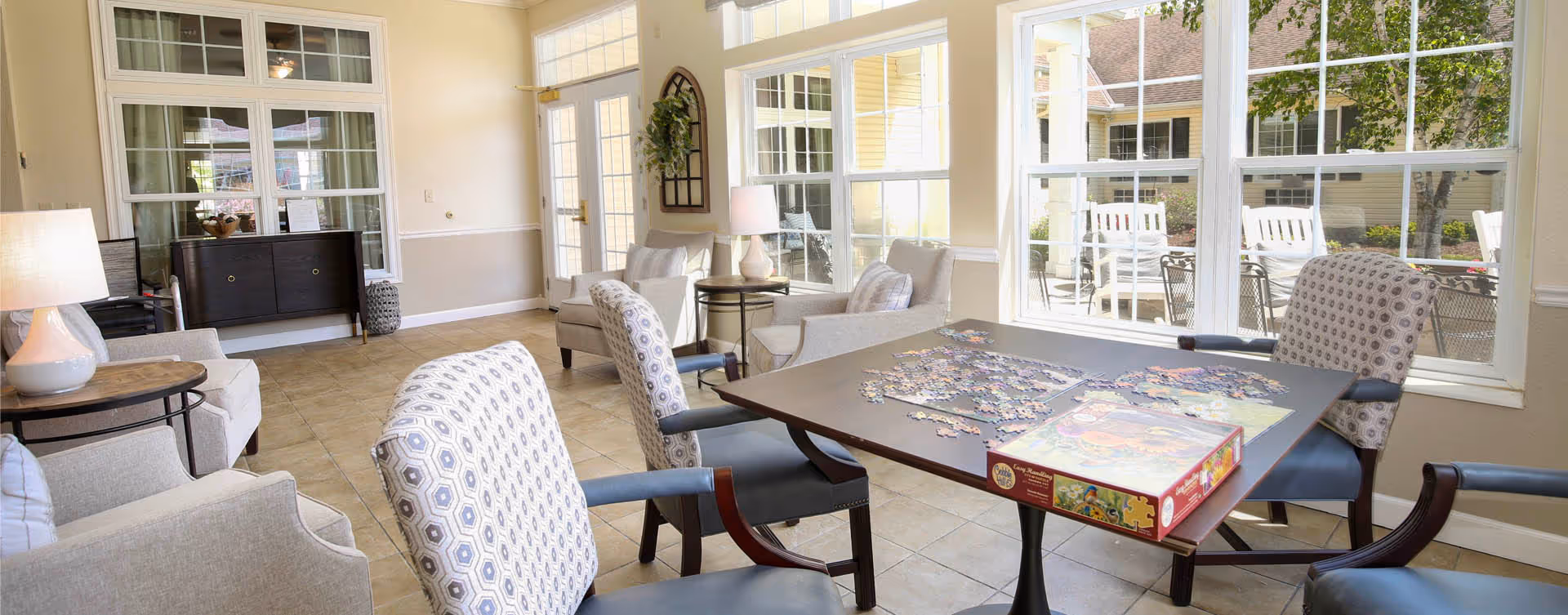 Bright communal lounge with armchairs around a table where a jigsaw puzzle is being assembled and large windows overlooking an outdoor patio.