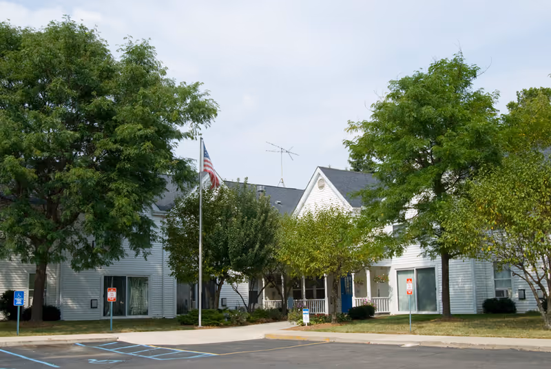 White two-story senior living building front with trees, an American flag, and a small parking area.