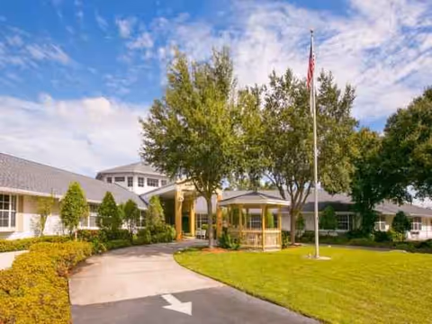 Senior living facility exterior with curved driveway, flagpole, trees, and covered entrance.