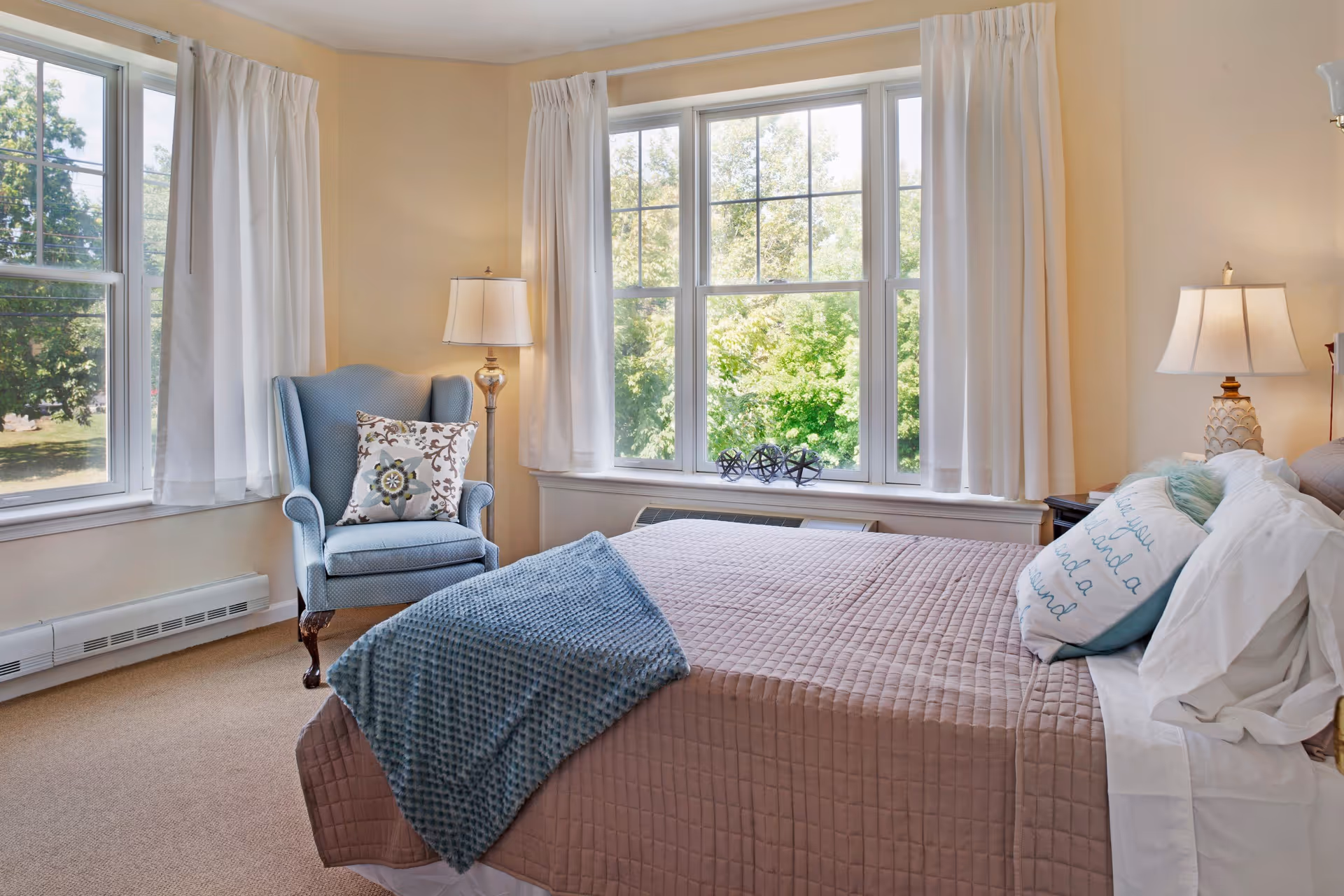Sunlit bedroom with a quilted bed, decorative pillows, a blue armchair and large windows overlooking greenery.