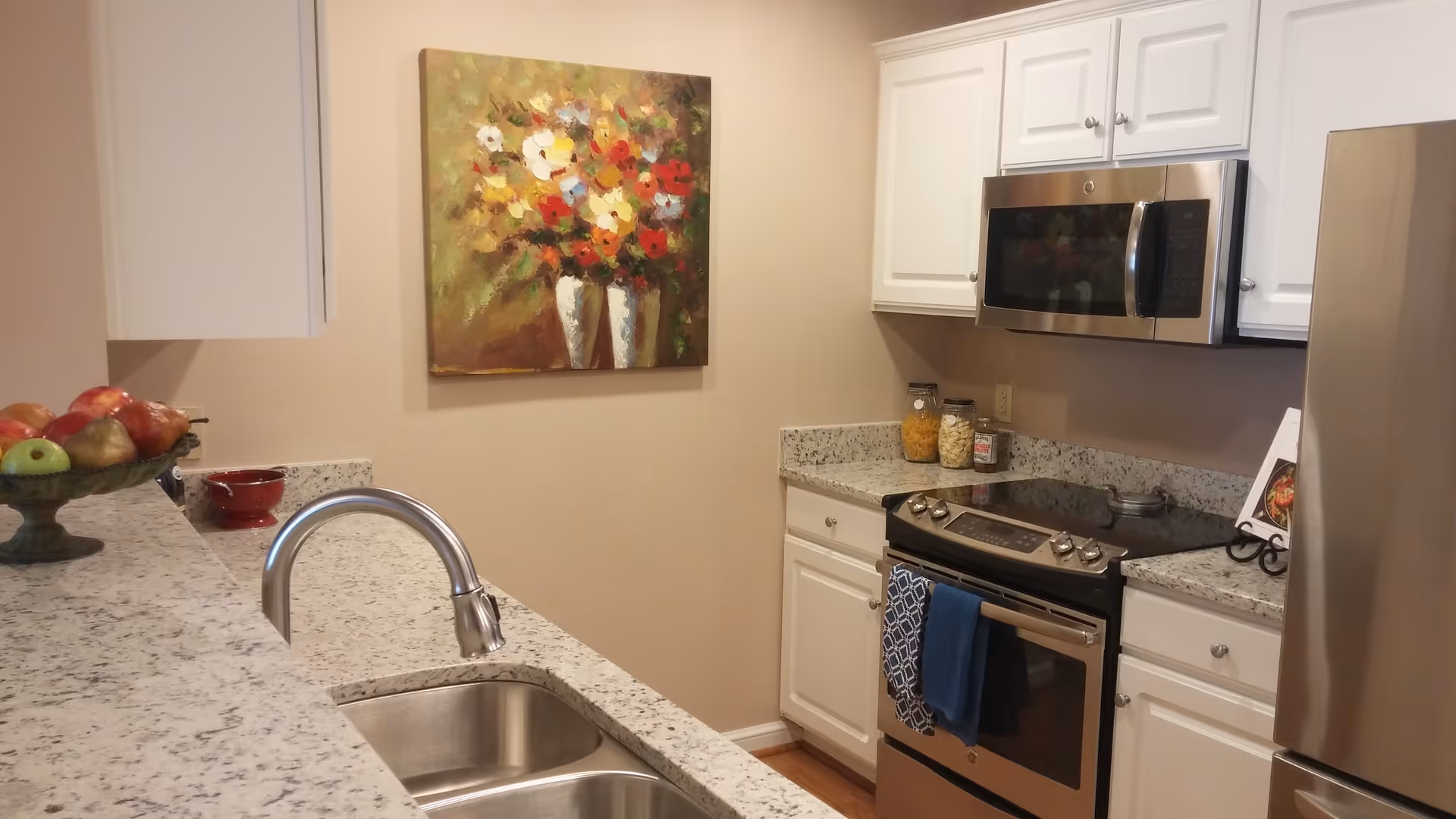 A modern kitchen with white cabinets, granite countertops, a stainless steel sink with a curved faucet, a stainless steel microwave above a stove with a blue towel hanging on the handle, and a stainless steel refrigerator. There is a colorful floral painting on the beige wall and a bowl of fruit on the counter.