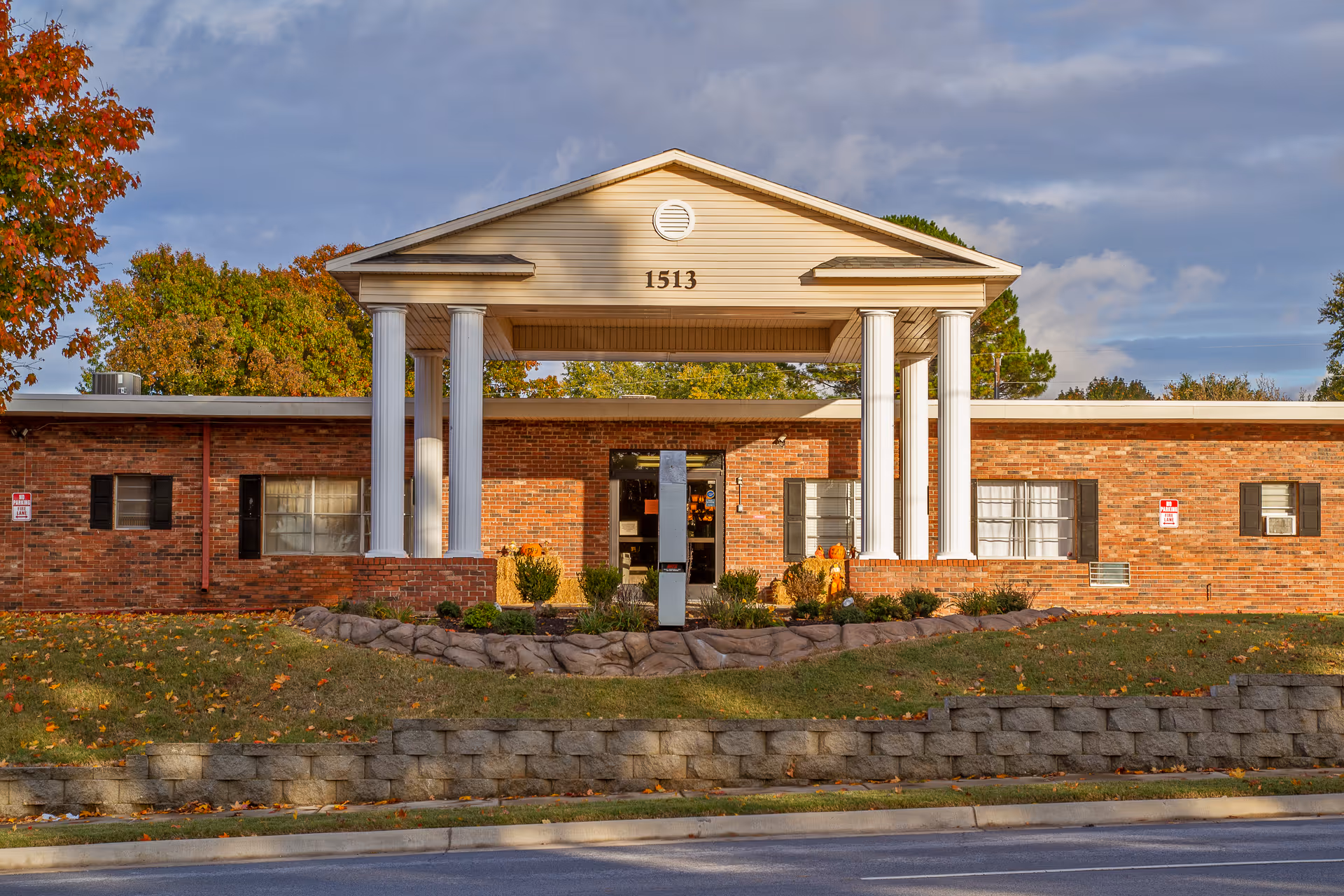 Front exterior view of a single-story brick building with white columns supporting a triangular pediment above the entrance. The building number 1513 is displayed on the pediment. There are windows with black shutters on either side of the entrance, and landscaping with bushes and a stone retaining wall in front. Trees with autumn foliage are visible in the background under a partly cloudy sky.