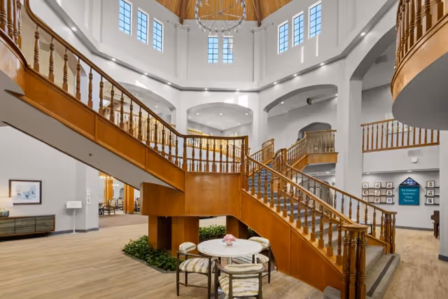 Spacious interior of a senior living community featuring a grand wooden staircase with railings leading to an upper level. The area is well-lit with natural light coming through multiple windows near the ceiling. Below the staircase is a small round table with four cushioned chairs and a flower centerpiece. The walls are painted white, and the floor is light wood. There are framed pictures and a sign on the walls in the background.