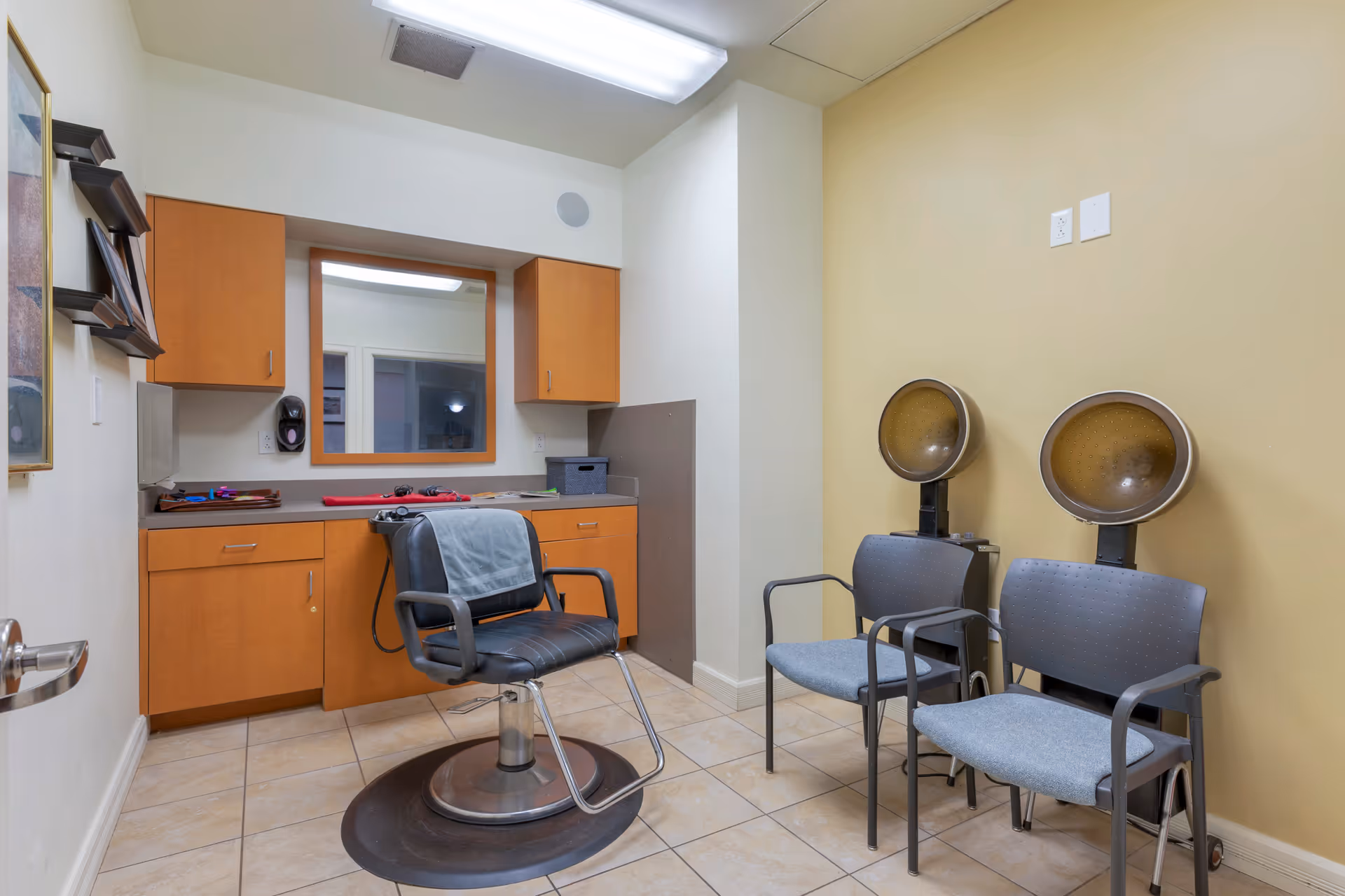 Interior view of a small salon room with a black salon chair in front of a counter with wooden cabinets and a large mirror. Two gray chairs with attached hair dryers are positioned against a beige wall on the right side. The floor is tiled and the room is well-lit with ceiling lights.