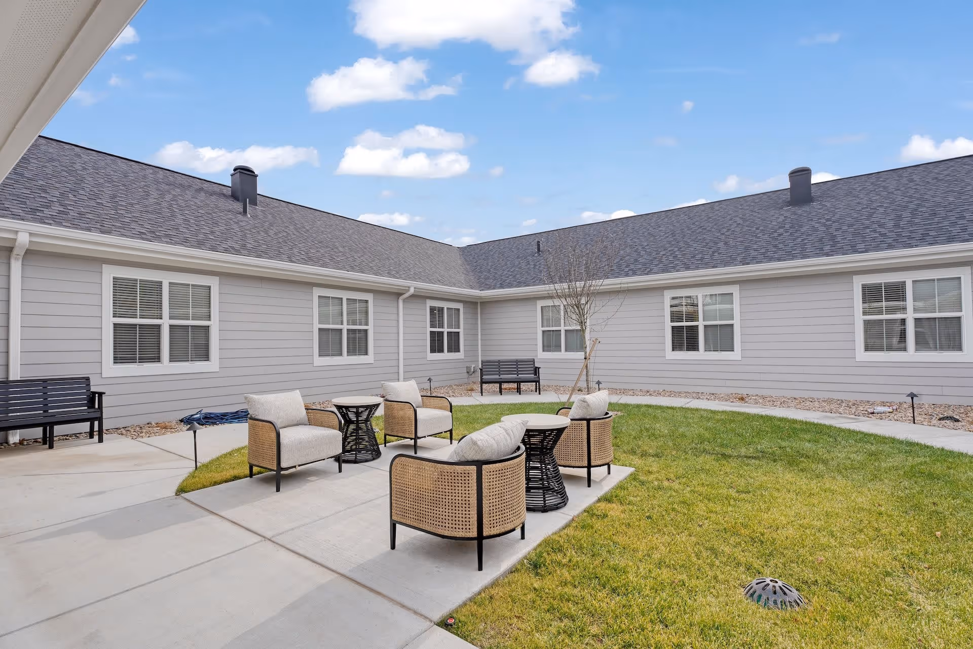 Outdoor courtyard area at Cedarhurst Senior Living of Salina featuring a concrete patio with four cushioned wicker chairs and two small round tables, surrounded by green grass and bordered by a single-story building with multiple windows under a partly cloudy blue sky.