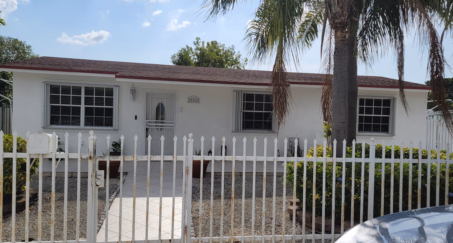 White single-story house with a palm tree, front yard and a white metal picket fence in front.