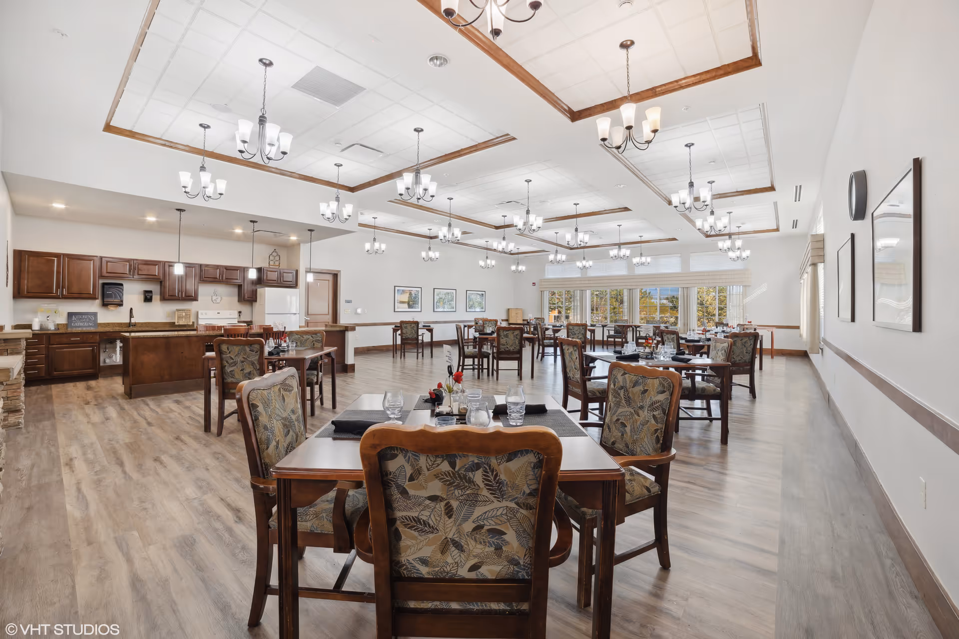 A spacious dining room with multiple wooden tables and chairs featuring floral upholstery. The room has a high ceiling with recessed lighting and several chandeliers. There is a kitchen area with wooden cabinets, a white refrigerator, and a stove along one wall. Large windows allow natural light to fill the room, and framed artwork decorates the walls.