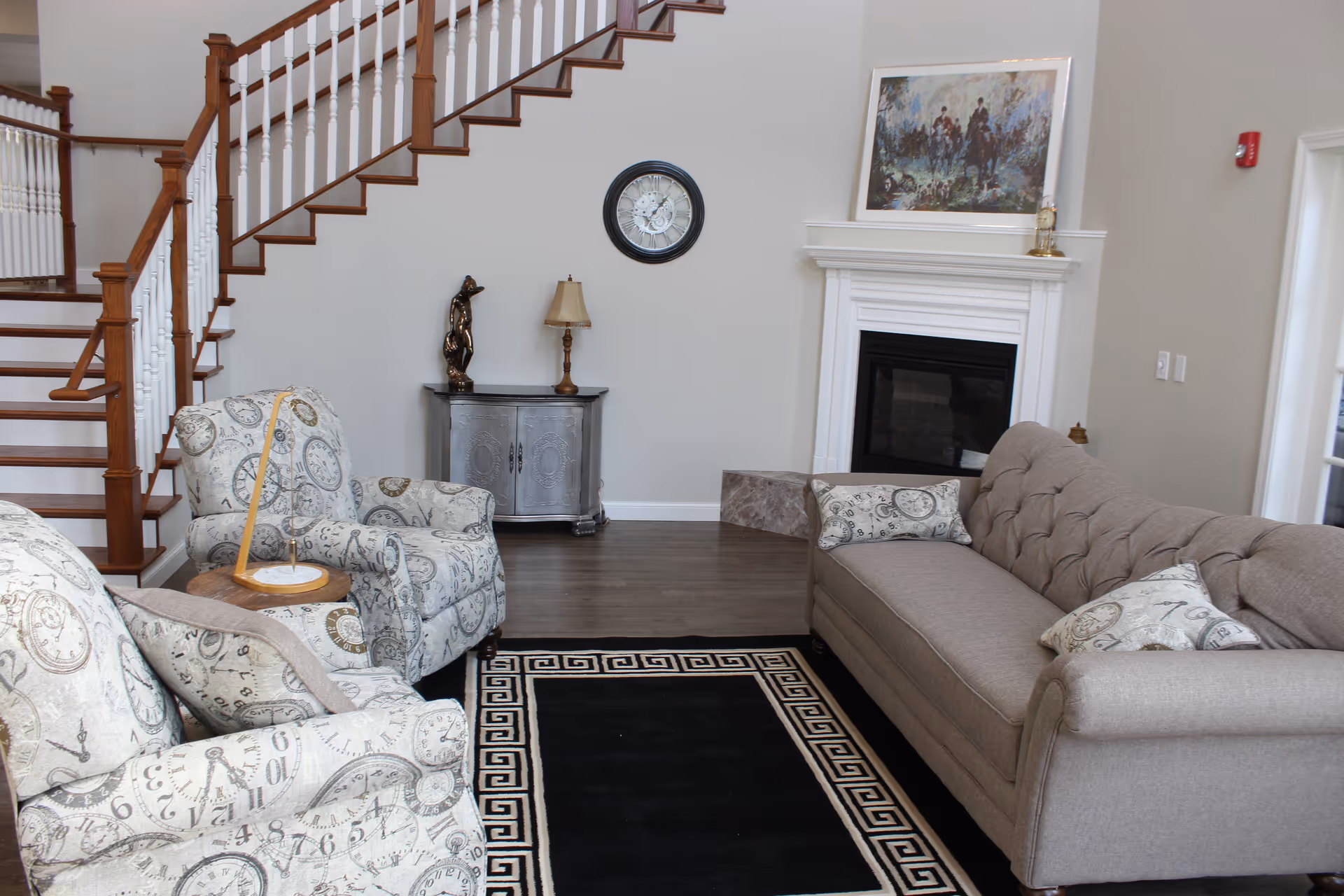 Living room with patterned armchairs, a tufted sofa, fireplace, staircase, and a black area rug.