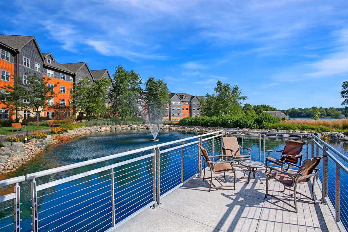 Outdoor patio area with four chairs and a small round table overlooking a pond with a water fountain. In the background, there are multi-story residential buildings, trees, and a clear blue sky.