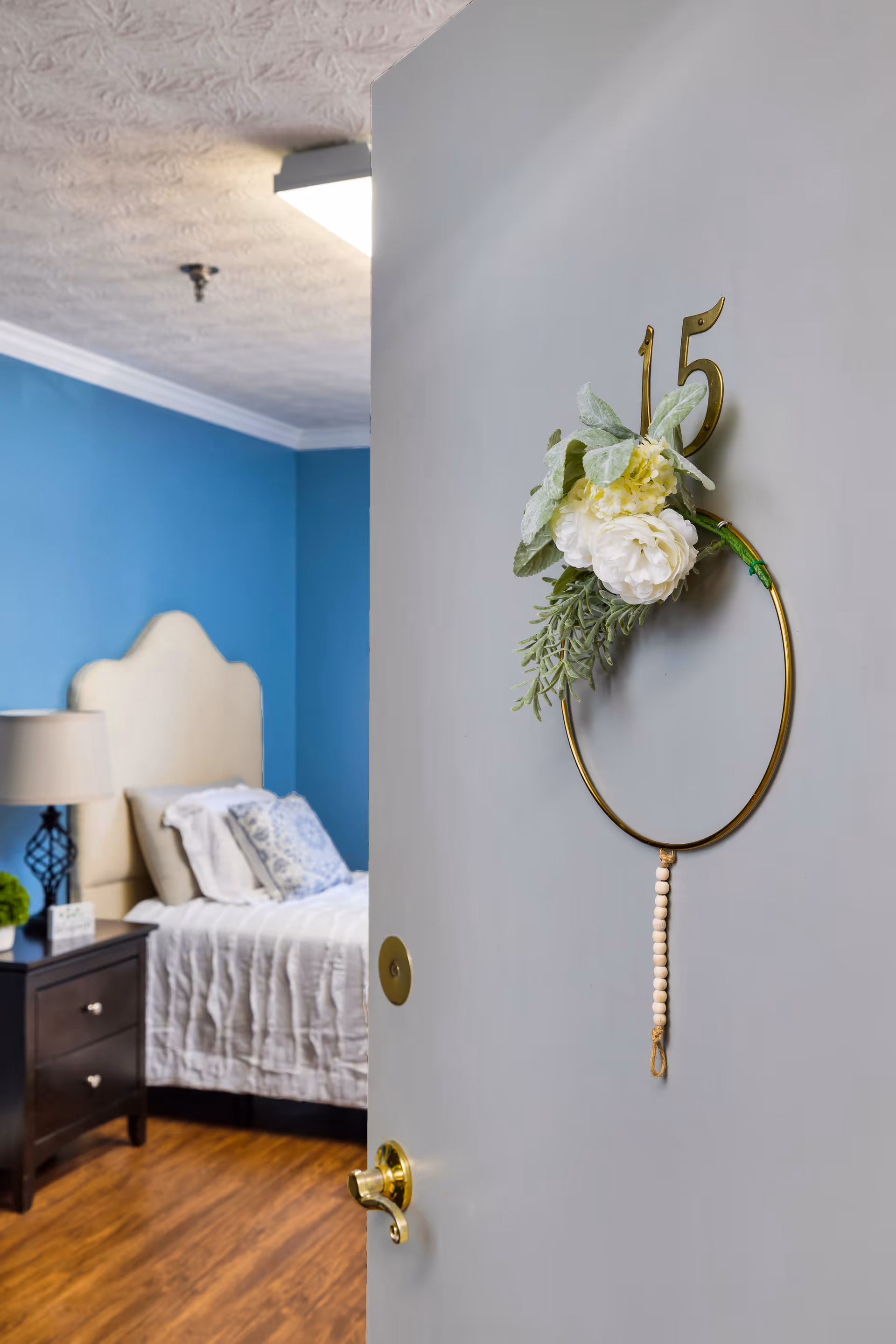 View through a partially open door into a bedroom with a blue accent wall, a bed with white bedding and patterned pillows, a nightstand with a lamp and a small plant. The door has a decorative gold ring with white flowers and greenery hanging on it, along with the number 15.