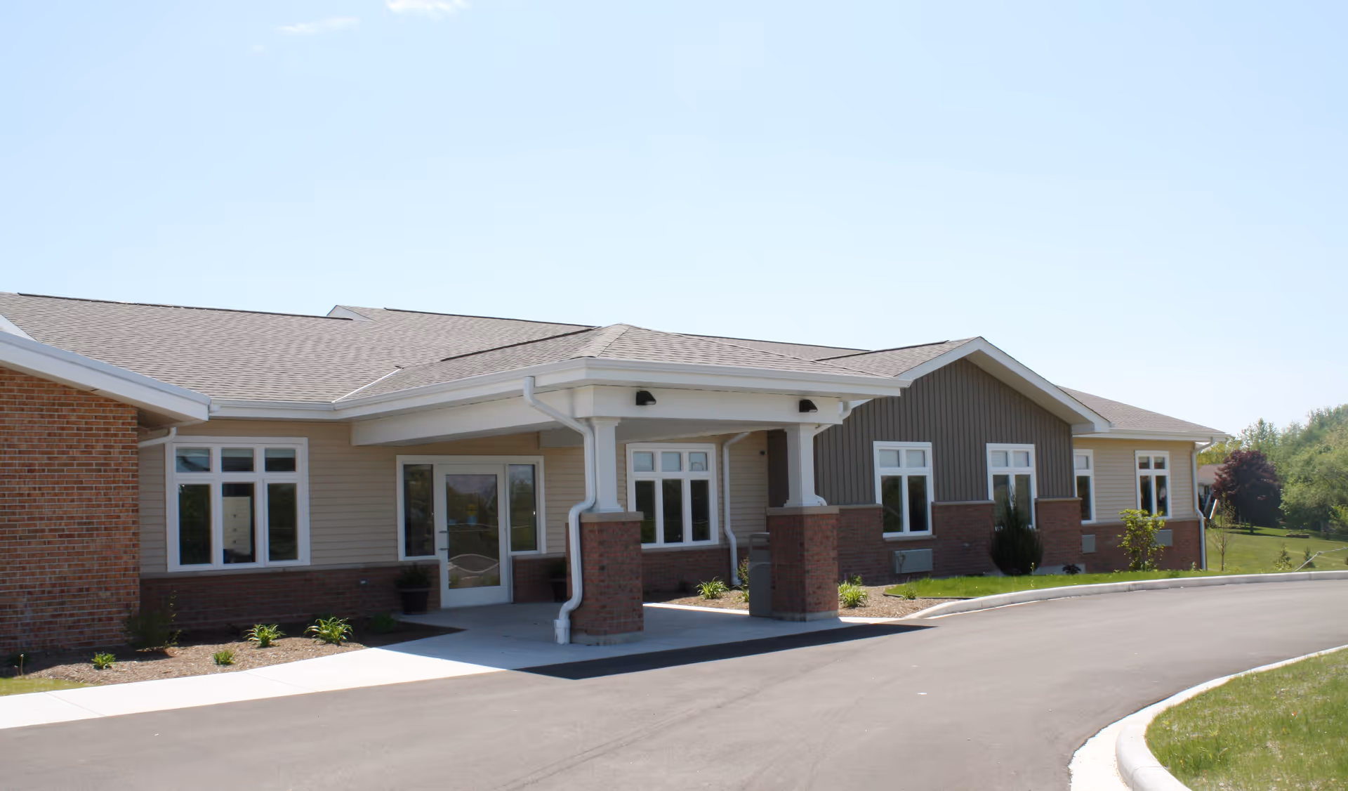 Exterior view of a single-story senior living facility building with a covered entrance, multiple windows, and a paved driveway surrounded by grass and landscaping under a clear blue sky.
