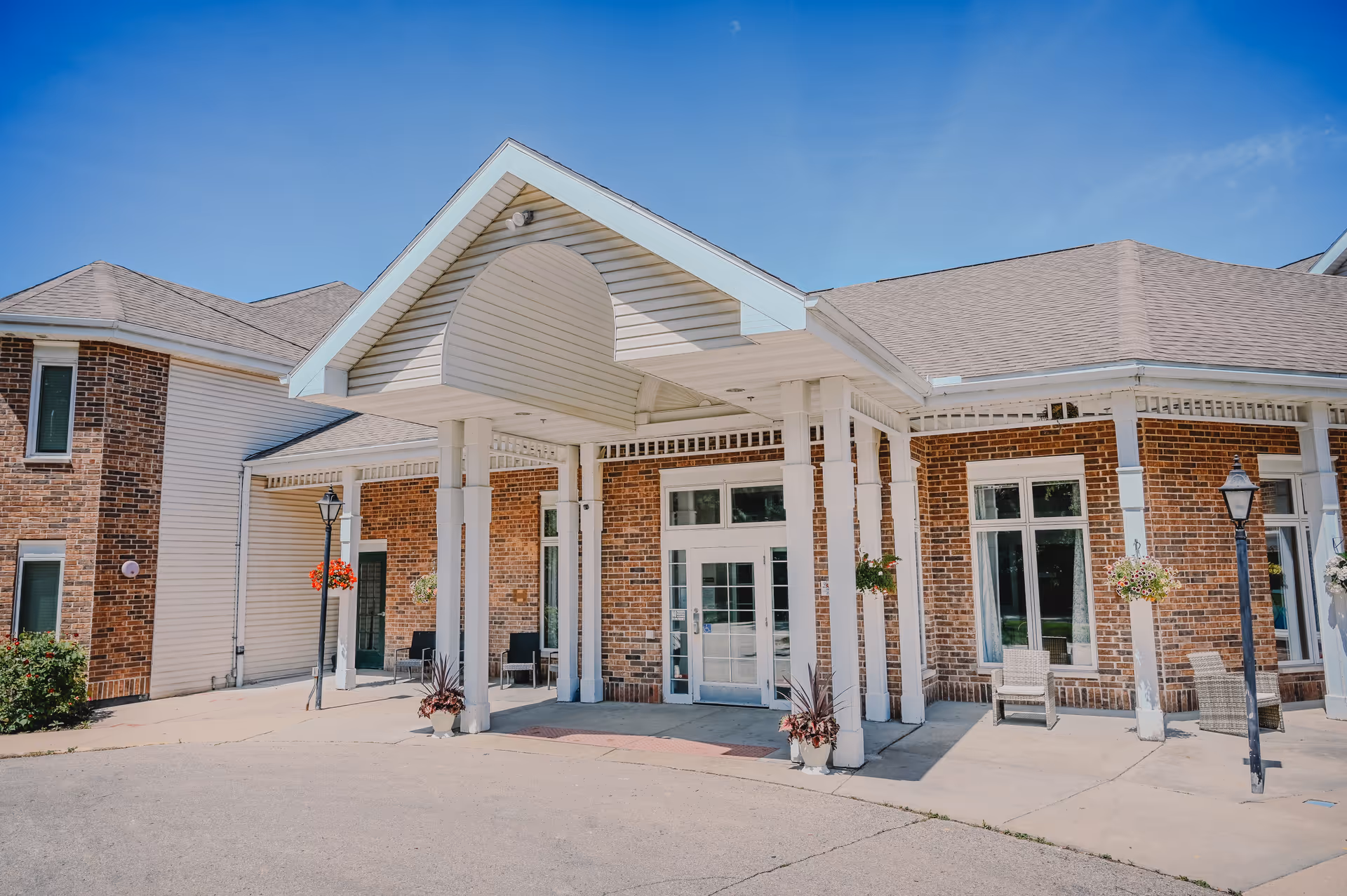 Front entrance of a senior living facility with a covered drop-off area supported by white pillars, brick and siding exterior walls, several windows, outdoor chairs, hanging flower baskets, and two black lamp posts under a clear blue sky.