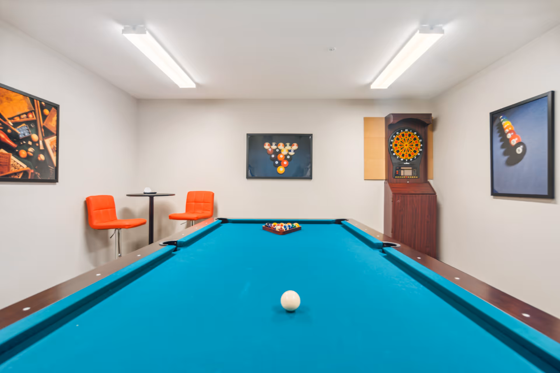 A game room with a pool table in the center, a dartboard on the right wall, and three framed pictures of billiard balls on the walls. There are two orange chairs and a small round table against the left wall. The room has white walls and ceiling lights.