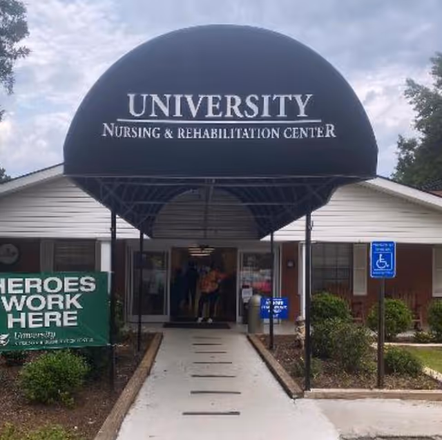 Entrance of University Nursing & Rehabilitation Center with a black canopy displaying the facility name. There is a concrete walkway leading to the entrance, a green sign on the left that reads 'HEROES WORK HERE,' and a blue handicap parking sign on the right. Some people are visible inside the building through the glass doors.