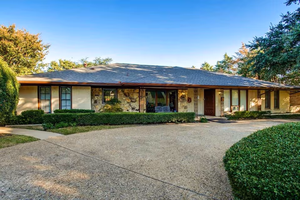Single-story building with a stone and beige exterior, surrounded by neatly trimmed bushes and trees under a clear blue sky. The building has multiple windows and a covered entrance with a bench nearby.