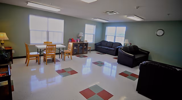 A spacious common area with large windows letting in natural light. The room has a tiled floor with red and green square patterns. There are two black leather sofas and one black leather armchair arranged near the windows. A wooden table with four chairs is positioned near two windows on the left side. A wooden cabinet with a lamp and decorative items is against the wall between the windows. A clock is mounted on the wall above the sofas.